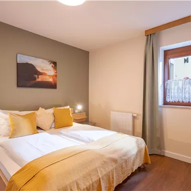 Ein gemütliches Schlafzimmer mit einem Doppelbett und gelben Kissen. Das Zimmer hat warme Farben und ein Fenster mit Vorhängen.