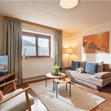 Ein gemütliches Wohnzimmer mit einem Sofa, einem Couchtisch und einem Fernseher. Es hat große Fenster und helle, freundliche Farben.