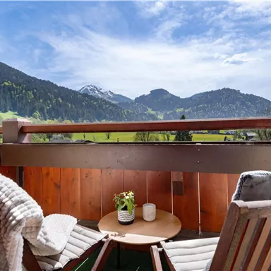Ein gemütlicher Balkon mit Holzstuhl und Tisch. Im Hintergrund sind grüne Hügel und Berge unter einem blauen Himmel zu sehen.