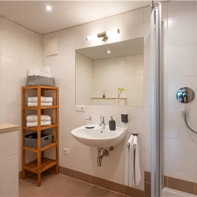 Ein modernes Badezimmer mit einer Dusche, einem Waschbecken und gefalteten Handtüchern. Ein Holzregal mit zusätzlichen Handtüchern ergänzt den Raum.