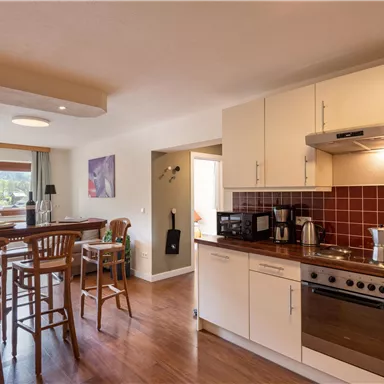 Eine moderne Küche mit Holzfußboden und hellen Möbeln. Im Hintergrund steht ein Tisch mit Stühlen und großen Fenstern, die viel Tageslicht hereinlassen.