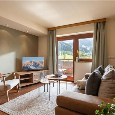 Ein gemütliches Wohnzimmer mit einem Sofa, einem Fernseher und einem Tisch. Große Fenster bieten einen Blick auf die grünen Berge.