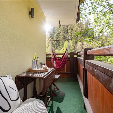 Ein gemütlicher Balkon mit einem Holztisch und einer Hängematte. Im Hintergrund sind Bäume zu sehen und das Licht der Sonne strahlt auf die Fläche.