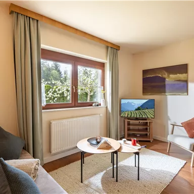 Ein gemütlicher Wohnraum mit einem hellen Sofa und einem kleinen Tisch. Große Fenster lassen viel Licht herein und bieten einen Blick ins Grüne.