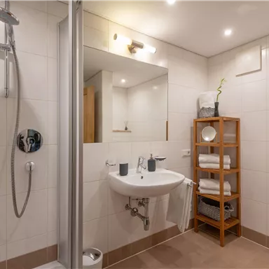 Ein modernes Badezimmer mit Dusche, Waschbecken und Spiegel. Es gibt einen Holzregal mit Handtüchern und Dekoration.