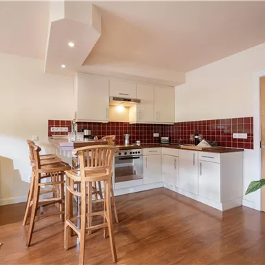 Eine moderne Küche mit Holzfußboden und weißen Schränken. Im Hintergrund stehen Barstühle und eine gemütliche Couch.