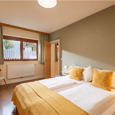 Ein gemütliches Schlafzimmer mit einem Doppelbett und bunten Kissen. Der Raum ist hell und hat große Fenster mit Blick nach draußen.