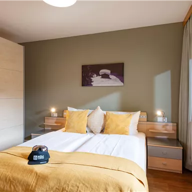 Ein modernes Schlafzimmer mit einem Doppelbett und gelben Kissen. Der Raum hat helle Möbel und ein großes Fenster mit Blick nach draußen.