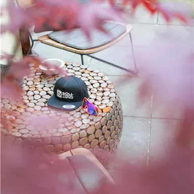 Ein schwarzer Hut liegt auf einem runden Tisch aus Holzscheiten. Im Hintergrund sind ein Stuhl und grüne Pflanzen zu sehen.