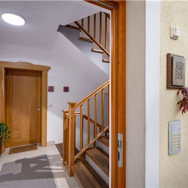 Ein einladender Flur mit Holztreppe und einem warmen Lichtquellen. Links befindet sich eine Eingangstür und rechts ein dekorativer Pflanzenbereich.
