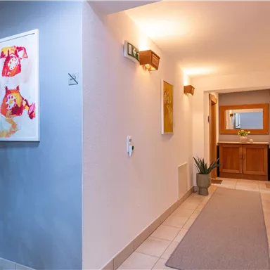 Ein heller Flur mit modernen Kunstwerken an den Wänden. Auf der rechten Seite befindet sich ein Holzschrank mit Pflanzen und einem Spiegel.