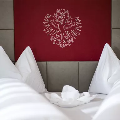 Ein gemütliches Zimmer mit einem großen Bett und vielen weichen Kissen. An der Wand ist ein stilisiertes Wappen abgebildet.