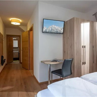 A bright room with two single beds, a desk, and a TV on the wall. The wood paneling and curtains create a cozy atmosphere.