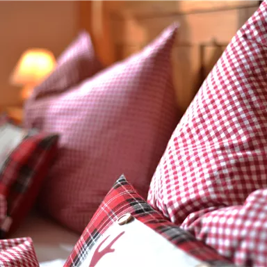 Kissen in roten Karomustern liegen auf einem Bett. Im Hintergrund ist eine sanfte Lichtquelle sichtbar.
