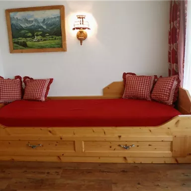 Ein gemütliches Holzbett mit roter Bettwäsche und karierten Kissen. Im Hintergrund hängt ein Landschaftsgemälde.