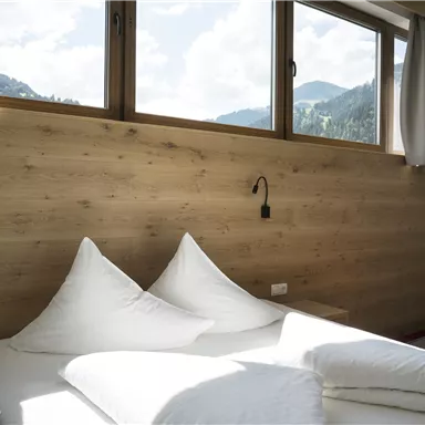 Ein helles Schlafzimmer mit Holzpaneelen und großen Fenstern. Zwei große, weiße Kissen liegen auf dem Bett, während die schönen Berglandschaften sichtbar sind.