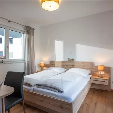 A modern bedroom with a double bed and wooden furniture. The room is bright and has large windows for natural light.