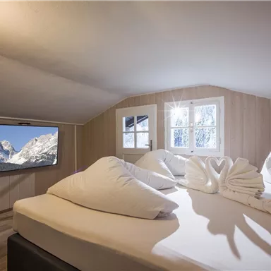 Ein gemütliches Schlafzimmer mit einem großen Bett und dekorativen Kissen. An der Wand hängt ein Fernseher, während helle Fenster den Raum erhellen.
