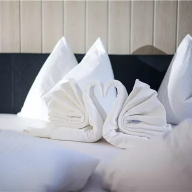 Ein Bett mit weißen Kissen und gefalteten Handtüchern in Form von Schwänen. Die Dekoration vermittelt eine romantische und einladende Atmosphäre.