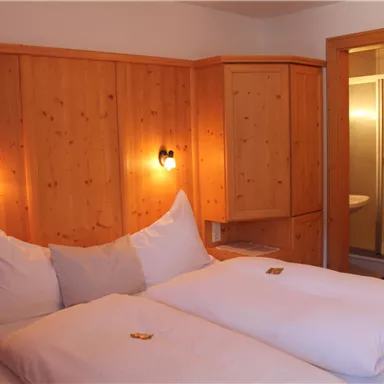 Ein gemütliches Schlafzimmer mit zwei Betten und Holzvertäfelungen. Im Hintergrund befindet sich ein modernes Badezimmer mit Dusche.