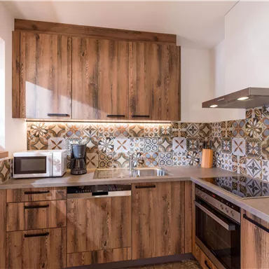 Eine moderne Küche mit Holzschränken und bunten Fliesen. Die Ausstattung umfasst eine Mikrowelle, einen Wasserkocher und einen Herd.