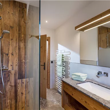 Ein modernes Badezimmer mit einer Glasdusche und hölzernen Akzenten. Die Einrichtung ist minimalistisch und elegant, mit einem Waschbecken aus Stein und Spiegeln.
