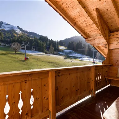 Ein schöner Balkon mit Holzverkleidung, der einen Blick auf grüne Wiesen und schneebedeckte Berge bietet. Die Sonnenstrahlen erhellen die Landschaft.