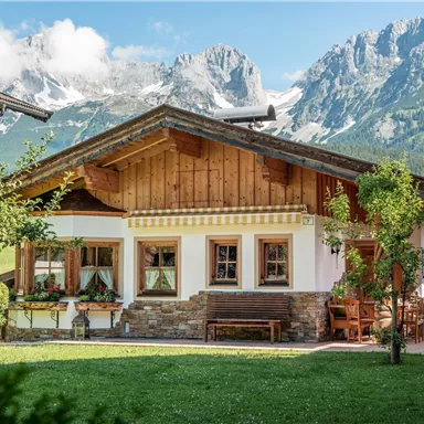 A cozy house in alpine style with a beautiful veranda. In the background, majestic mountains and a clear sky can be seen.