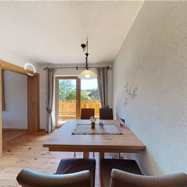A bright dining area with a wooden table and cozy chairs. In the background are windows overlooking the outside.