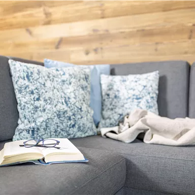 A cozy living room with a gray sofa, colorful cushions, and a blanket. On the table lies an open book and a pair of glasses.