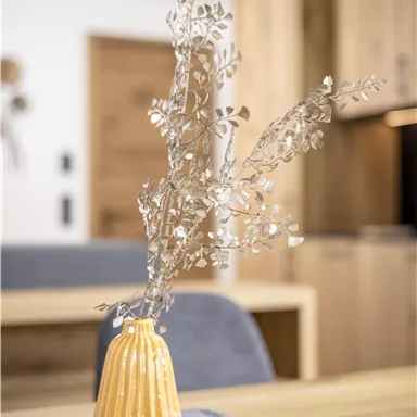 A yellow vase with silver branches stands on a wooden table. In the background, modern furniture and a neutral color palette are visible.