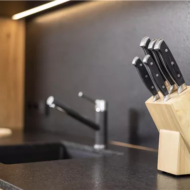 A modern kitchen environment with a wooden cutting block holding five sharp knives. In the background, a sink and a faucet are visible.