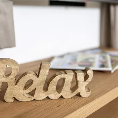 Ein dekoratives Holzschild mit der Aufschrift "Relax" steht auf einem Holztisch. Im Hintergrund liegen einige Zeitschriften.