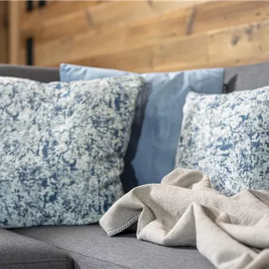 Ein gemütliches Sofa mit dekorativen Kissen in Blau- und Grautönen. Darauf liegt eine helle Decke, die eine einladende Atmosphäre schafft.
