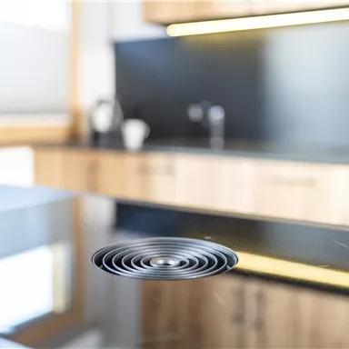 Eine moderne Küchentheke aus schwarzem Stein mit einem runden Dunstabzug. Im Hintergrund sind Holzschränke und Küchenutensilien sichtbar.