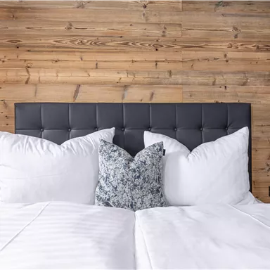 A cozy bedroom with a modern headboard and soft pillows. The wooden wall gives the room a warm atmosphere.