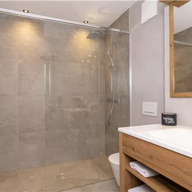 A modern bathroom with a large shower and gray tiles. The wash area has a wooden cabinet and a mirror.