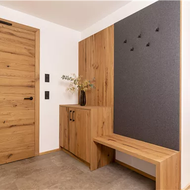 A modern entrance area with a wooden door and a stylish wooden bench. On the wall, there is a gray area with hooks and a vase with dried flowers.