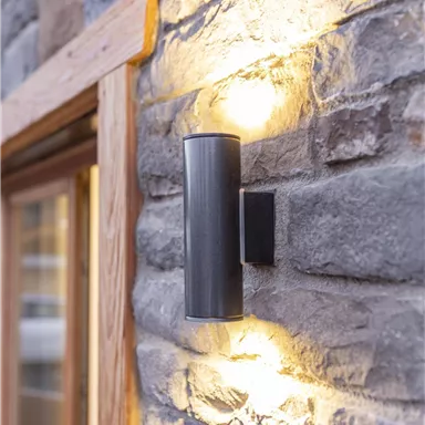 Eine moderne Außenlampe aus schwarzem Metall an einer steinernen Wand. Das Licht strahlt weich und schafft eine angenehme Atmosphäre.