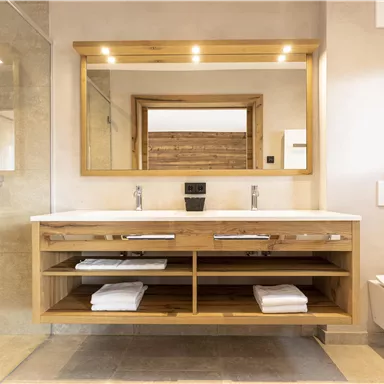 A modern bathroom with a large mirror wall and an elegant wooden washstand. The shower is made of glass and there are fresh towels on the open shelf.