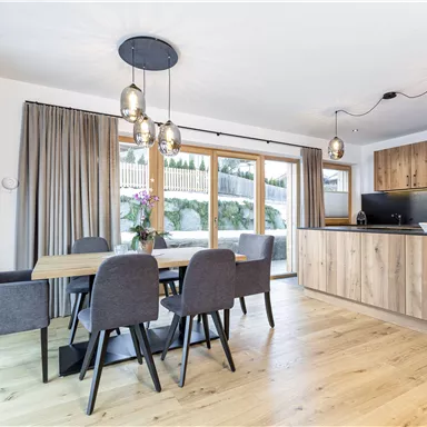 A modern kitchen with a dining area. Large windows let in plenty of light, and the furnishings consist of wood and gray chairs.