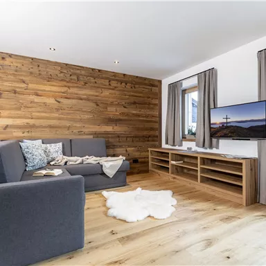 Ein modernes Wohnzimmer mit einer Holzvertäfelung und einem gemütlichen Sofa. Der Raum ist hell und einladend, ausgestattet mit einem Fernseher und einem Fenster mit Vorhängen.