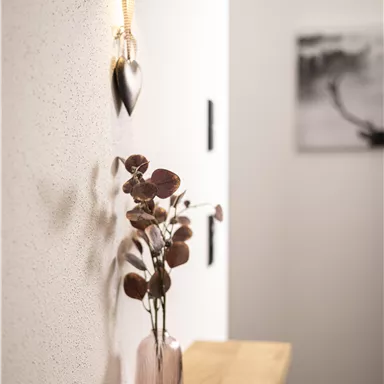 A stylish interior with a wooden console and a pink vase filled with dried branches. A modern lamp hangs on the wall along with a heart-shaped decorative piece.