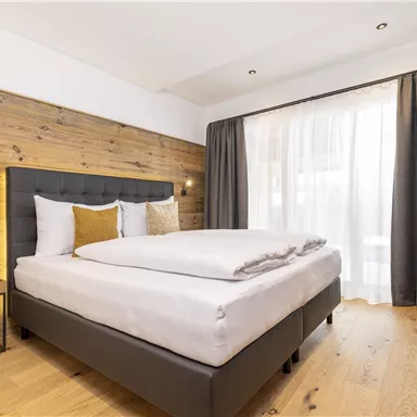 Ein modernes Schlafzimmer mit einem komfortablen Bett und Holzpaneelen an der Wand. Große Fenster lassen viel Tageslicht herein, und die Vorhänge sind elegant drapiert.