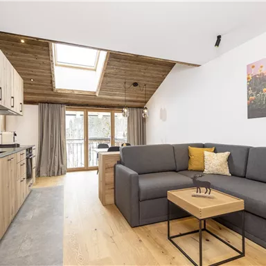 Ein modernes Wohnzimmer mit einer bequemen grauen Couch und einem kleinen Tisch. Die großen Fenster lassen viel Licht herein und es gibt Kunstwerke an der Wand.