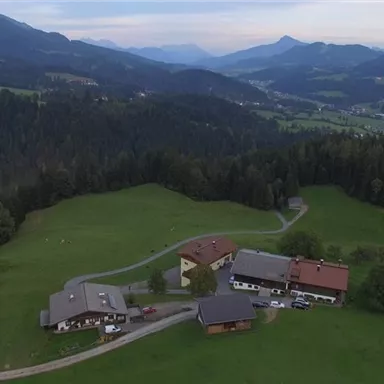 Eine malerische Landschaft mit mehreren Gebäuden auf einer Wiese. Im Hintergrund erstrecken sich sanfte Hügel und Berge unter einem bewölkten Himmel.