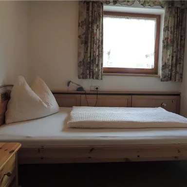 Ein einfaches Schlafzimmer mit einem Holzbett und zwei dekorativen Kissen. Der Raum hat ein Fenster mit Vorhängen und einen Holzschrank.