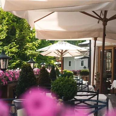 Ein eleganter Außenbereich eines Restaurants mit Tischen und Stühlen. Bunte Blumen und Sonnenschirme schaffen eine einladende Atmosphäre.