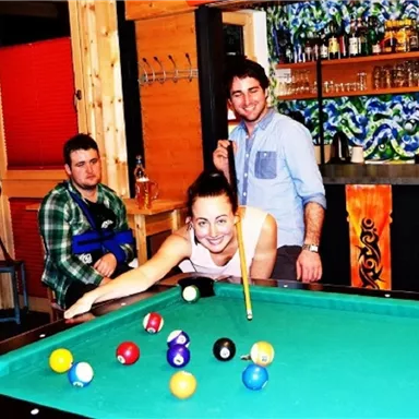 Eine gesellige Runde am Billardtisch mit Lächeln und Spaß. Im Hintergrund ist eine Bar mit bunten Flaschen zu sehen.