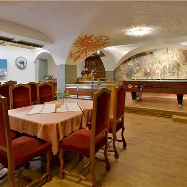 Ein gemütlicher Raum mit einem großen Tisch und roten Stühlen. Im Hintergrund befindet sich ein Billardtisch und eine Wandmalerei.